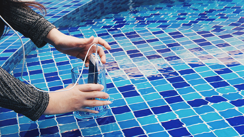 A person holding a glass half submerged in a swimming pool with a smartphone placed inside the glass 